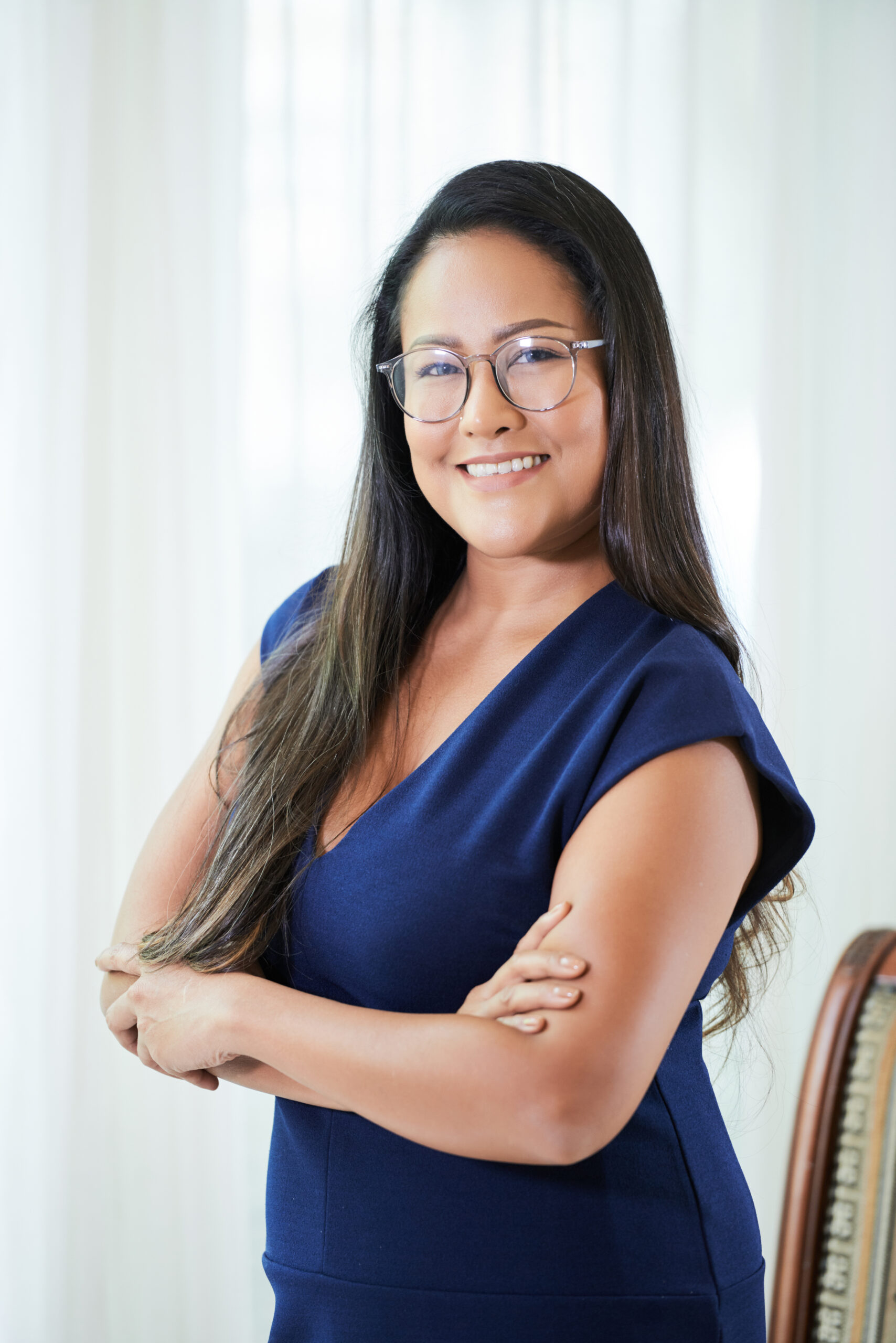 Cheerful adult Asian businesswoman in blue dress and glasses standing with hands crossed looking at camera